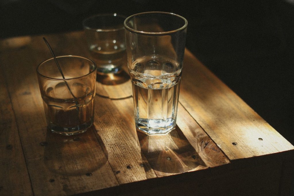 Водний баланс організму: скільки води пити щодня A warm, dramatic still life of drinking glasses casting shadows on a wooden table.