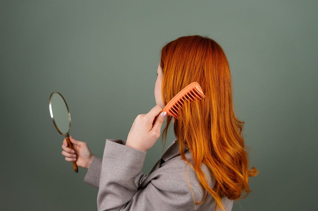 Як доглядати за волоссям вдома: практичні поради Red-haired woman using a comb while holding a mirror, focusing on hair care and beauty.