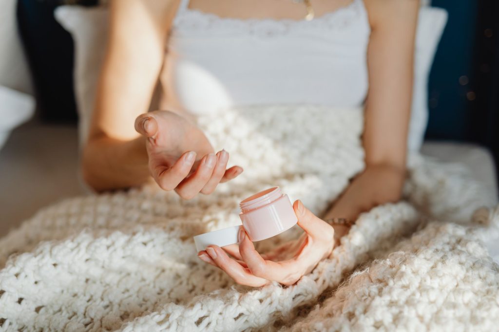 Догляд за шкірою взимку: захист від холоду й сухості A young woman uses moisturizer while wrapped in a cozy wool blanket indoors.