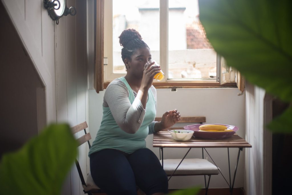 A woman sits and drinks juice by a window, enjoying a healthy break indoors.