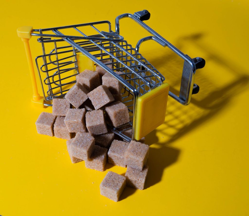 Менше цукру в раціоні: прості кроки до легкості Brown sugar cubes spilling from a small shopping cart on a vibrant yellow background.