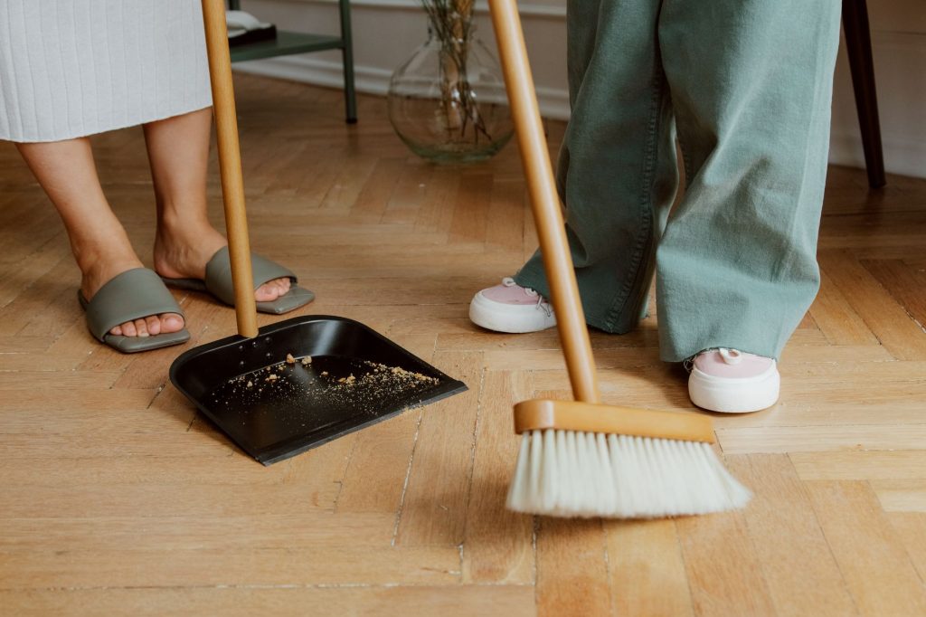 Як організувати прибирання квартири: система без хаосу Close-up of people cleaning wooden floor with broom and dustpan indoors.