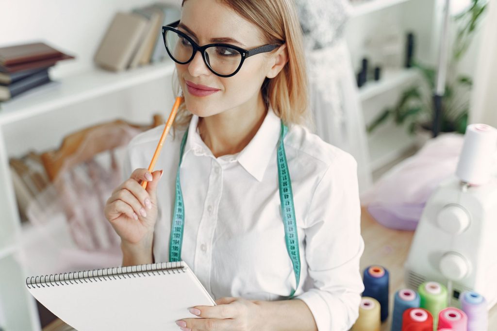 Creative fashion designer with glasses thinking in her studio with sketchpad and sewing machine.