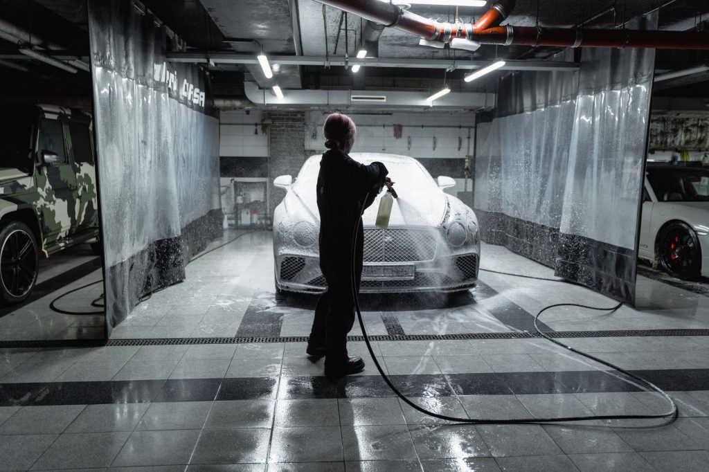 A person using a hose to spray a car with snow foam in an indoor carwash setting.